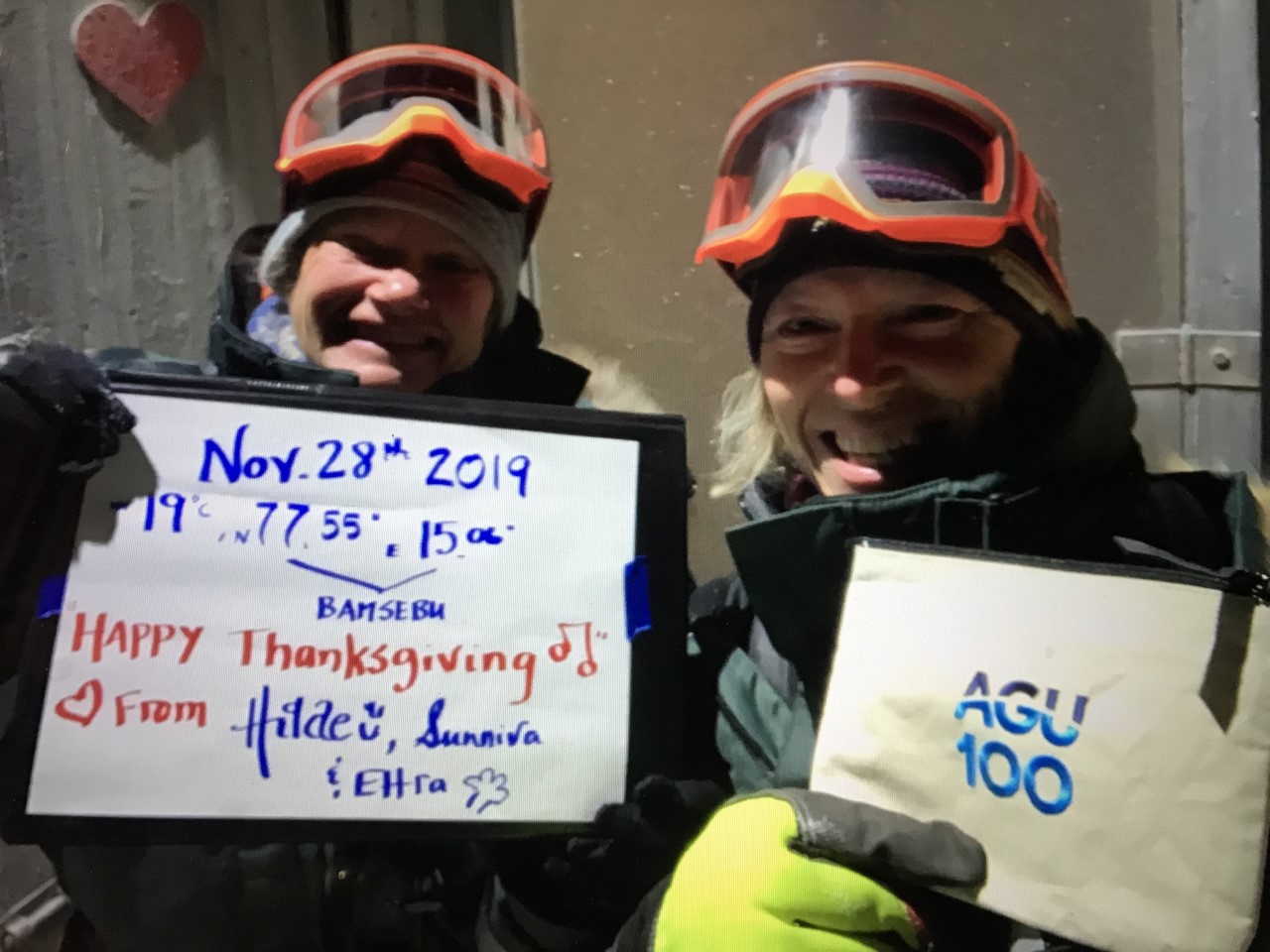 Two women in cold weather gear hold up sings that say 19 degrees Celsius, N 17.55, E 15.06, (Bamsebu), Happy Thanksgiving! (music notes) Love from Hilde, Sunniva, and Ettra" and "AGU 100."