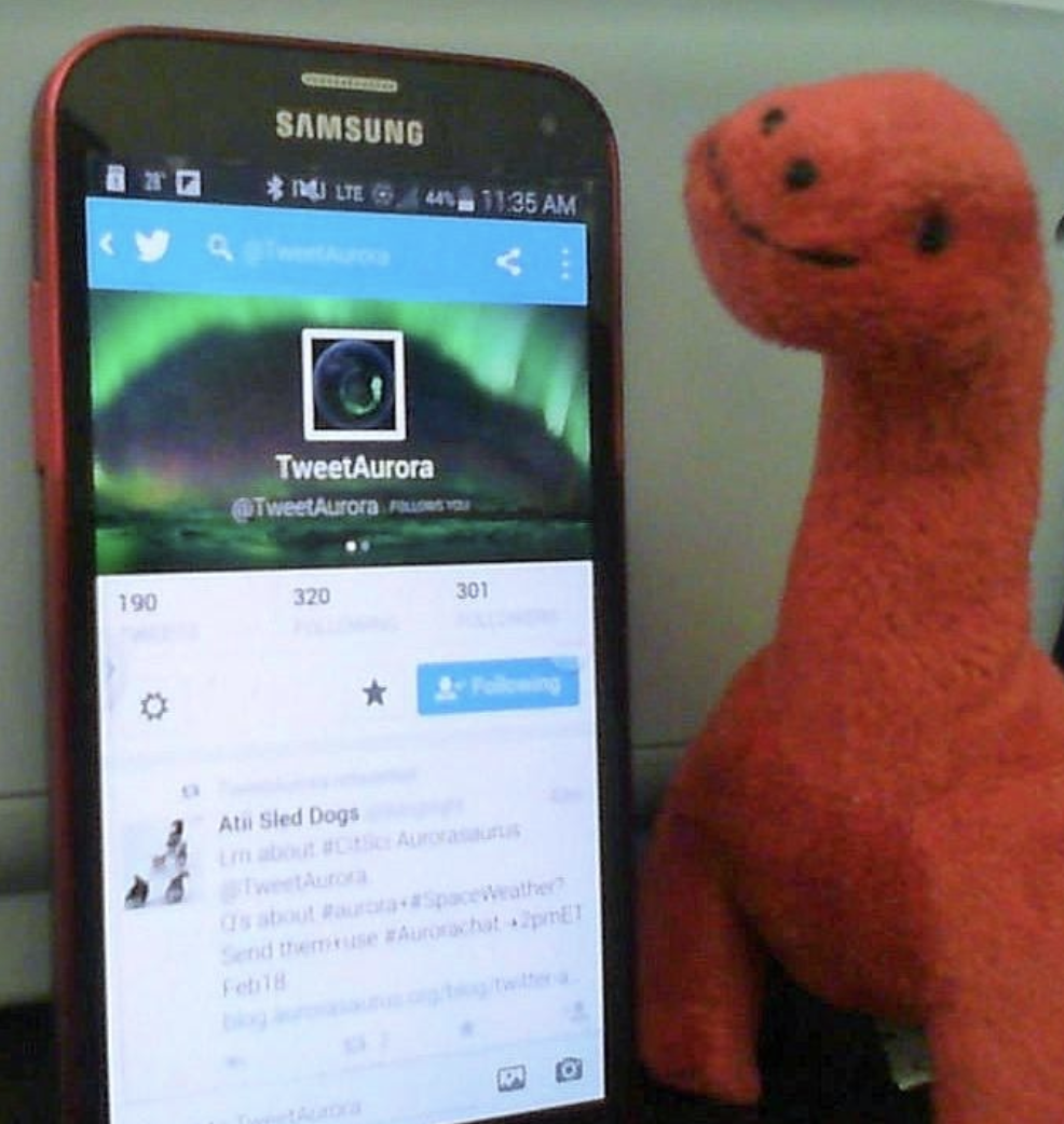 A smartphone on the Aurorasaurus Twitter sits next to a small red long-necked dinosaur plush