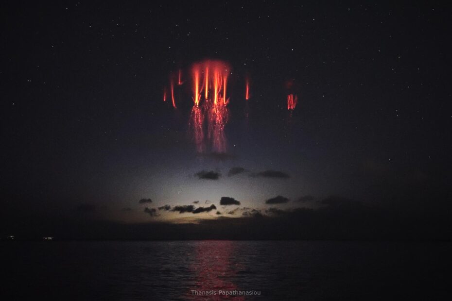 The center of a black background is illuminated. Clouds and a horizon are visible, but above them are sprites, appearing like bright red carrots with branching roots at the tips, buried in the night sky.