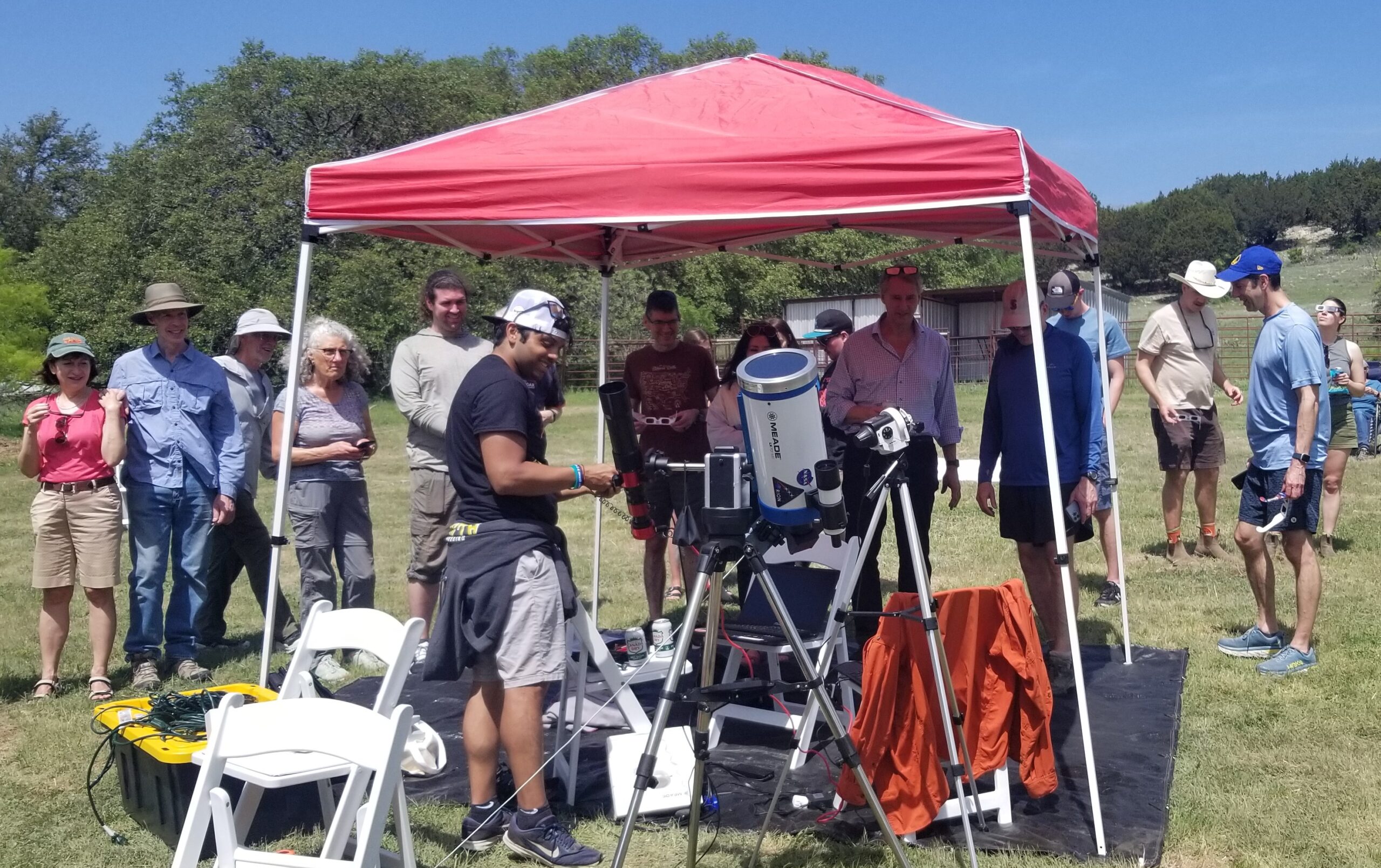 Under a red pop-up tent, a man gestures toward a solar telescope. He is surrounded by fascinated onlookers.