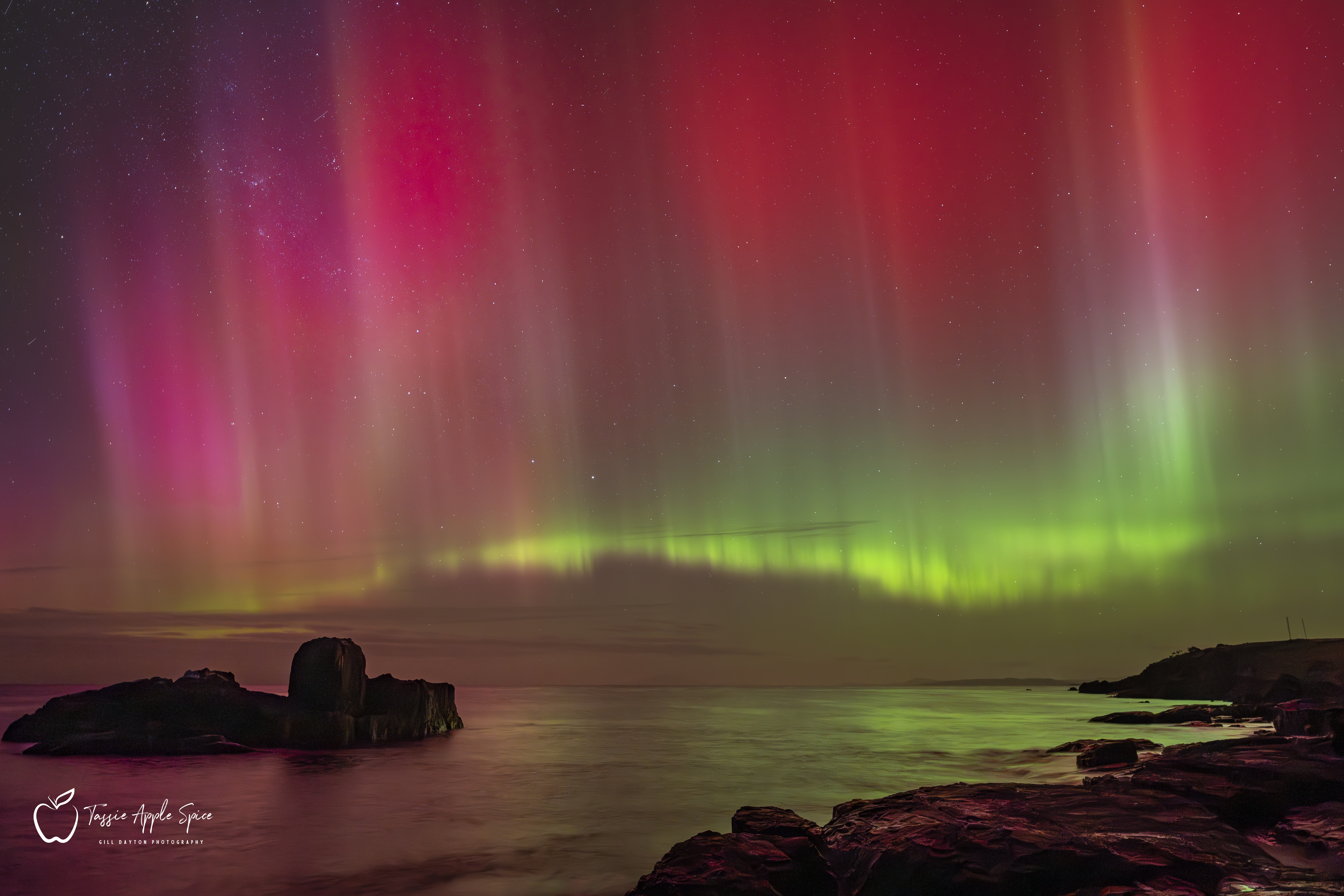 Above a rocky coastline, reflected in the sea, a ribbon of bright green light waves just above the horizon. Stretching above it are plumes of bright red light, with the stars shimmering through.