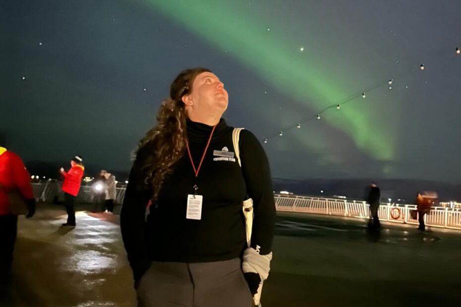 On the deck of a ship, a woman gazes up at a stripe of green aurora.