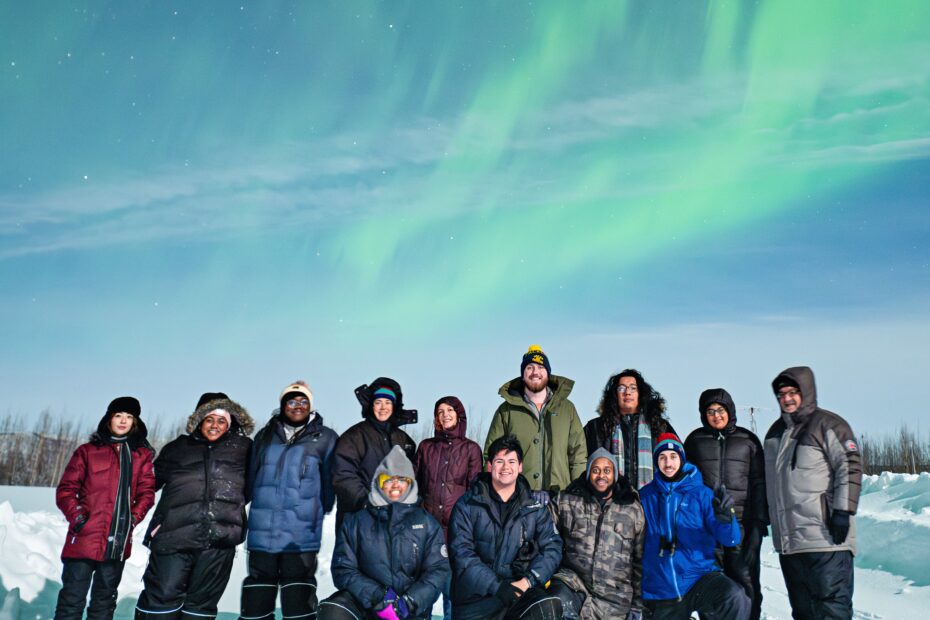 A group of people, bundled in cold weather gear, smile broadly beneath wispy green bands of aurora