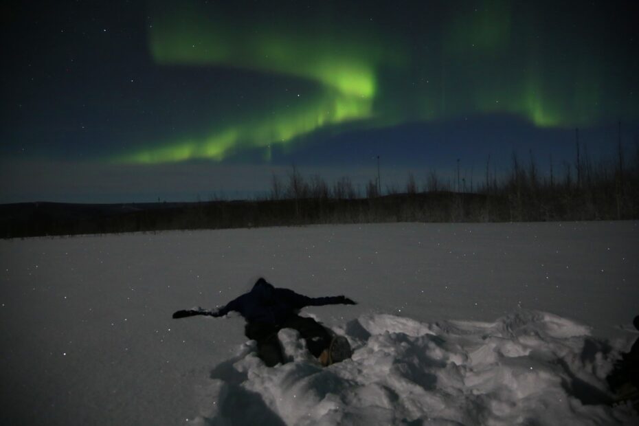 In a snowy landscape beneath bright green bands of aurora, a young person lies in the snow looking up at the lights