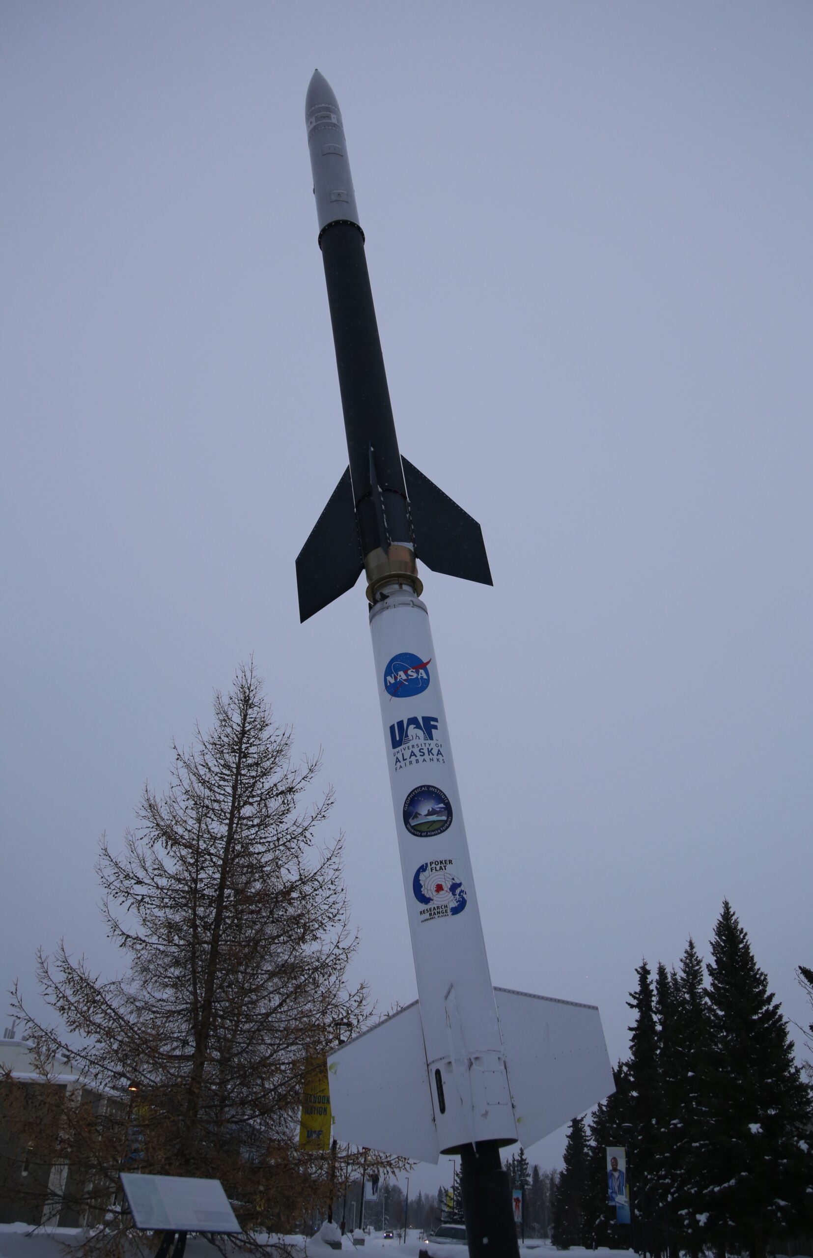 Above trees against a light grey sky, a rocket labeled NASA, UAF, UAFGI, and PFRR.