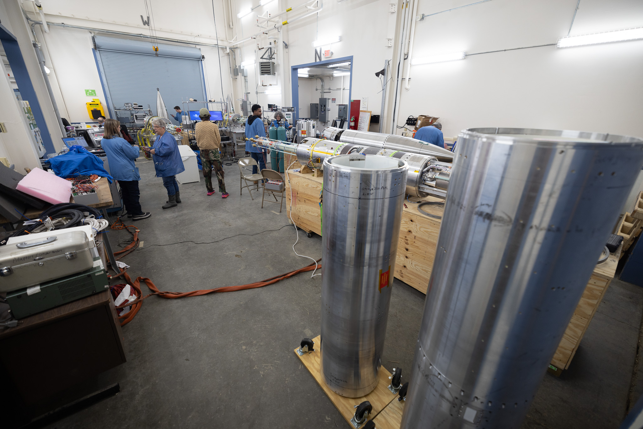 Behind a foreground of large silver-colored cylinders stood on end, a group of scientists talk and work.