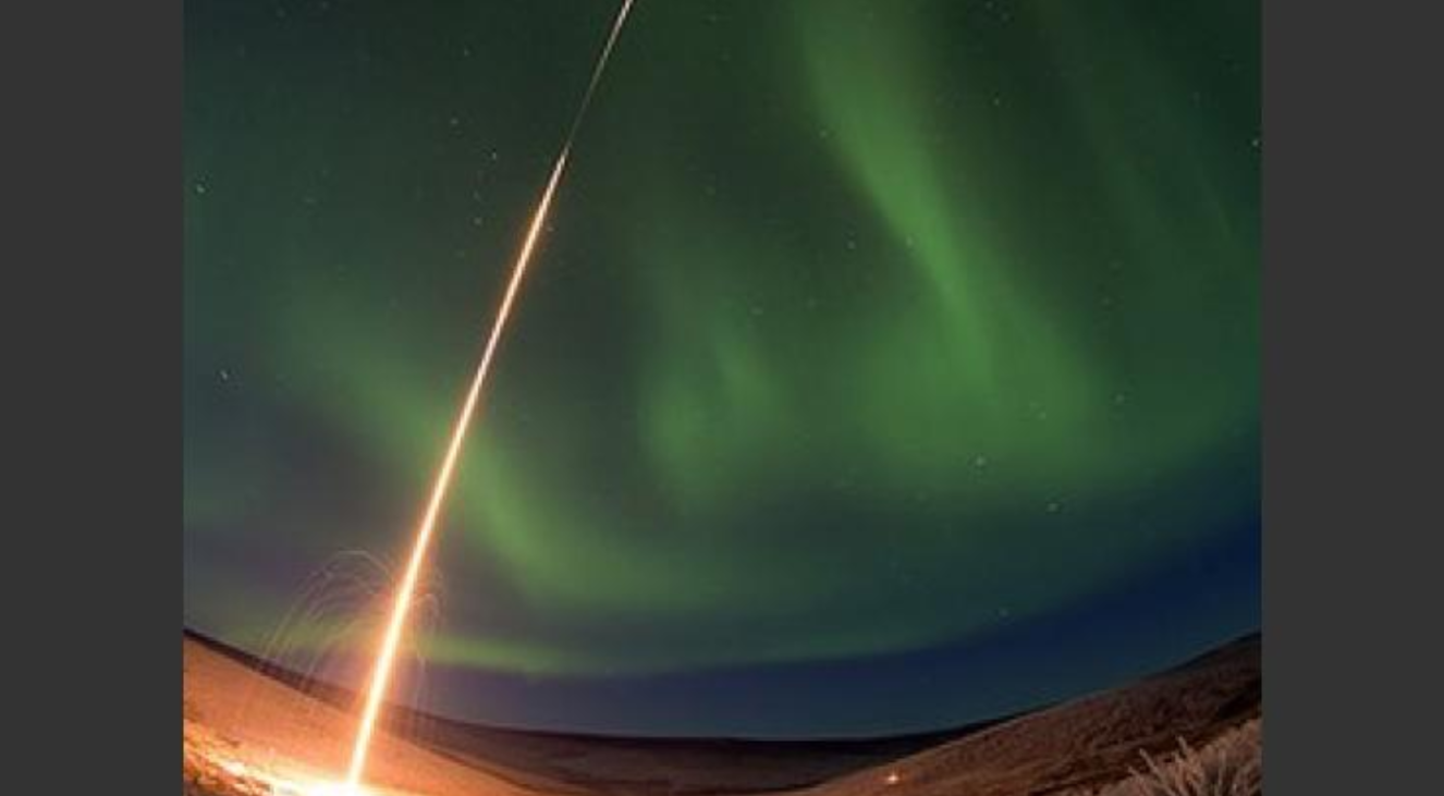 In a snowy landscape beneath a sky full of green aurora, an orange streak of light marks a scientific rocket launching into the aurora for study.