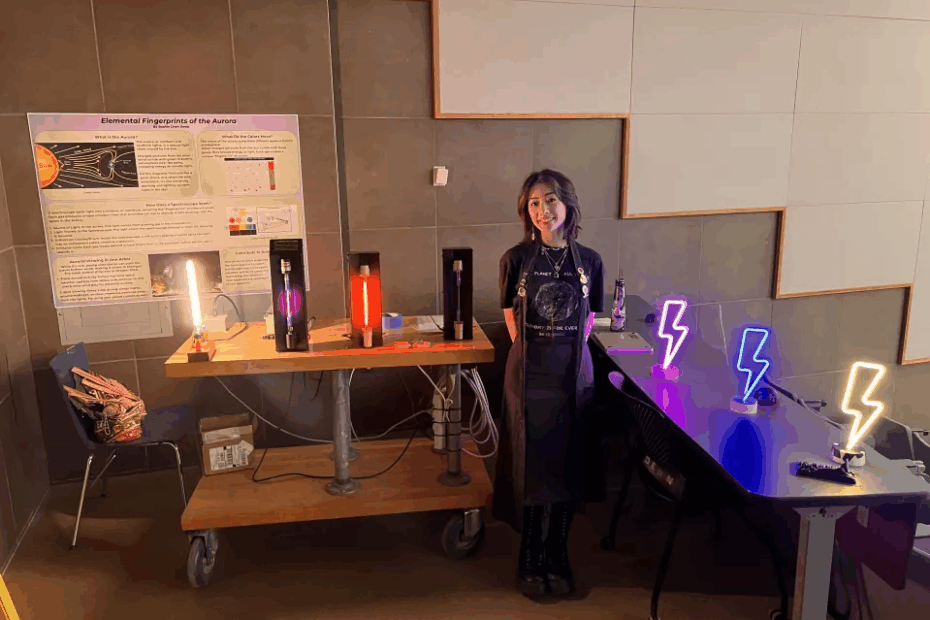 A young woman stands beside tables covered with neon lights in different colors. A scientific poster hangs in the background.