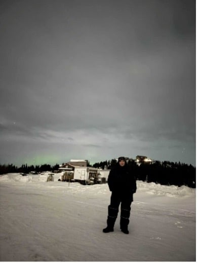 A young woman stands bundled up in a snowy, cloudy landscape with the faint glow of aurora on the horizon