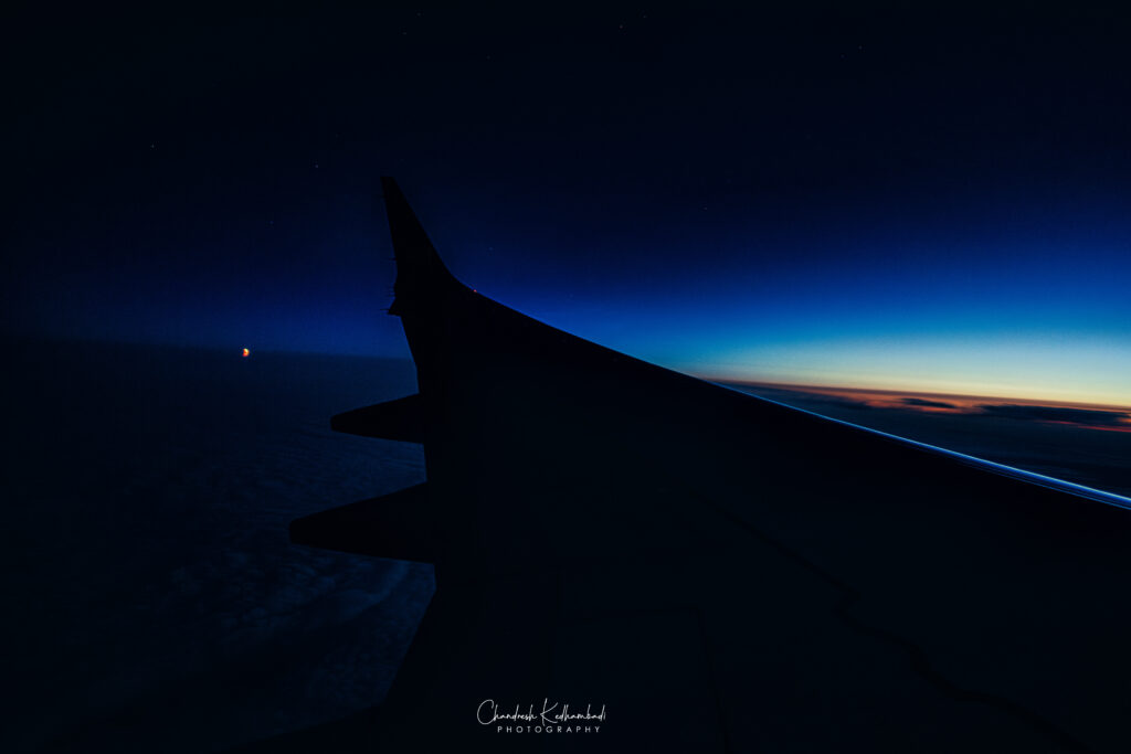 Over the wing of a plane moving toward the right of the photo, night stretches behind while dawn crests the horizon ahead.