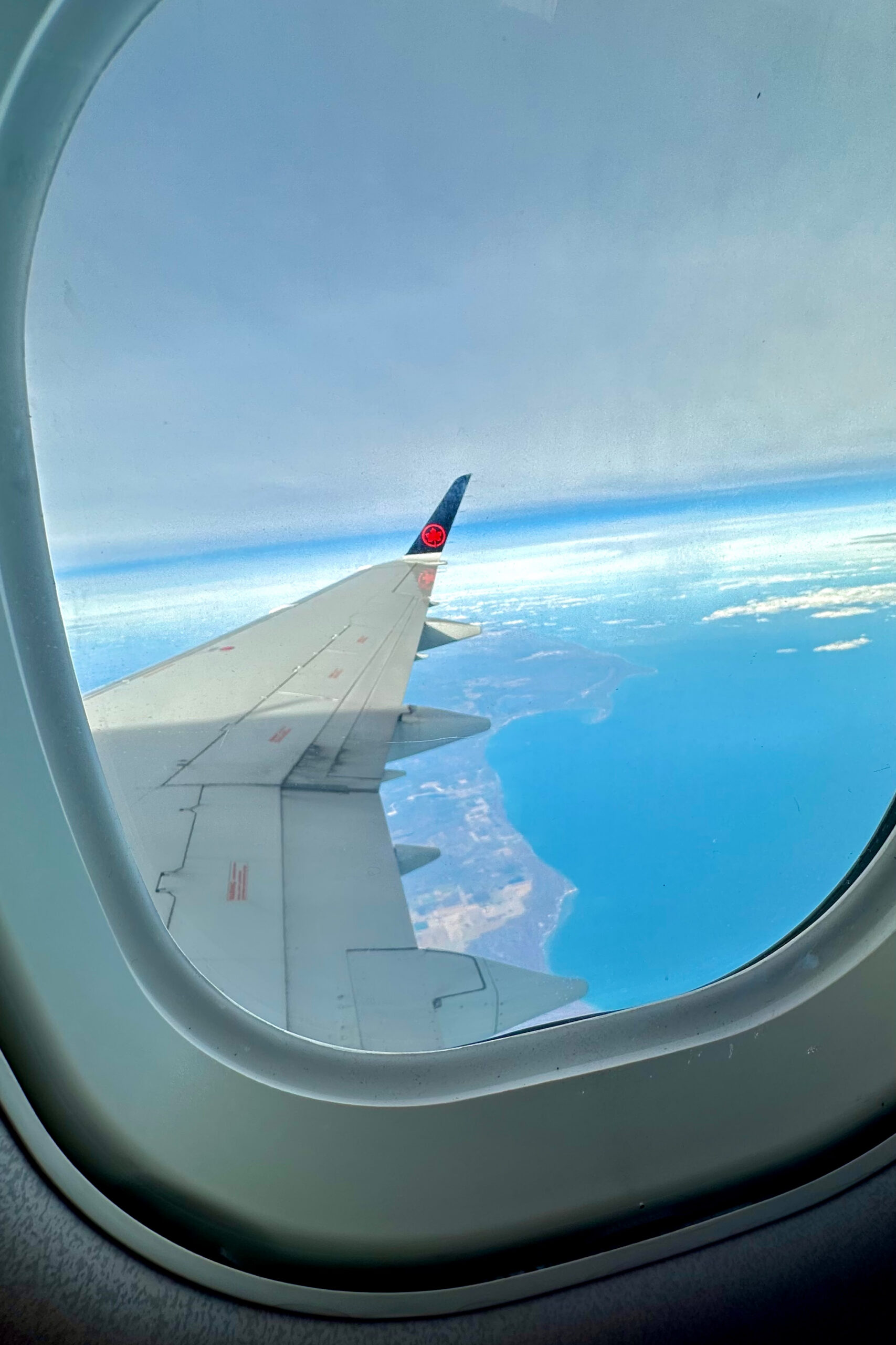 A daytime photo out a plane window shows a wing above a blue, cloudy landscape far below.