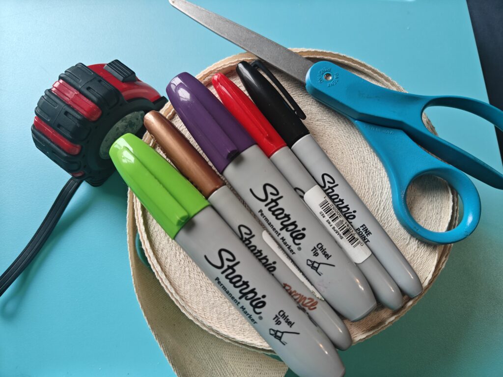 On top of a beige roll of fabric tape sit several colorful permanent markers and a pair of fabric scissors. Next to them is a hardware measuring tape.